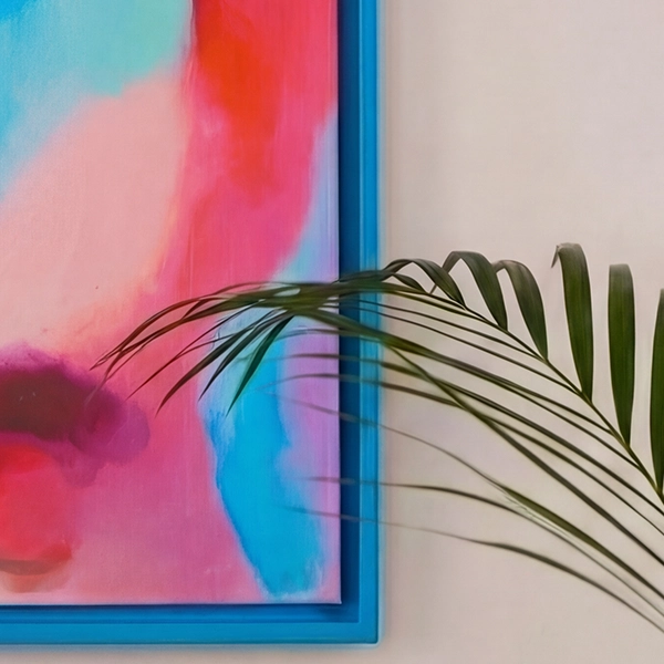 Cadre bleu avec toile rose et turquoise devant un mur beige clair. Une feuille de palmier décore le bord de l'image.